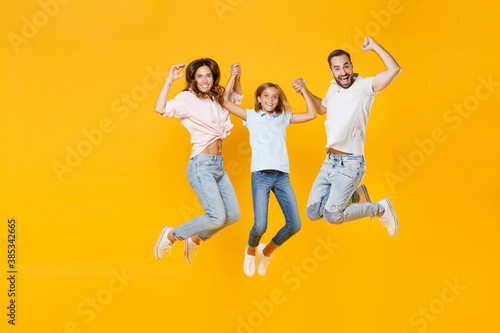 Full length portrait of cheerful young parents mom dad with child kid daughter teen girl in basic t-shirts jumping holding hands isolated on yellow background studio portrait. Family day concept.