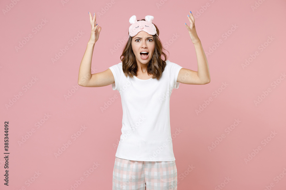 Shocked angry irritated young woman in white pajamas home wear sleep mask spreading hands screaming swearing while resting at home isolated on pink background studio portrait. Relax good mood concept. Shocked angry irritated young woman in white pajamas home wear sleep mask spreading hands screaming swearing while resting at home isolated on pink background studio portrait. Relax good mood concept.