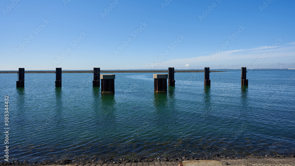 Dark wooden columns stand in the deep blue water, blue sky with white clouds, Dam in the foreground. Netherlands, Zeeland.