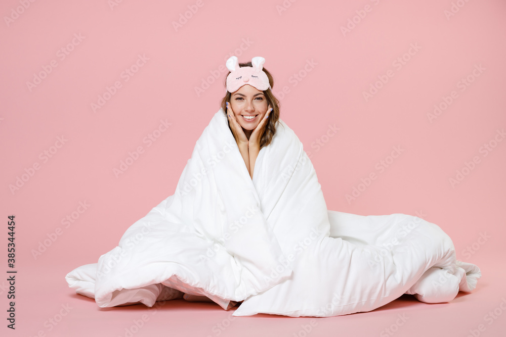 Smiling young woman in pajamas home wear sleep mask sitting wrapping in blanket put hands on cheeks looking camera resting at home isolated on pink background studio portrait. Relax good mood concept. Smiling young woman in pajamas home wear sleep mask sitting wrapping in blanket put hands on cheeks looking camera resting at home isolated on pink background studio portrait. Relax good mood concept.