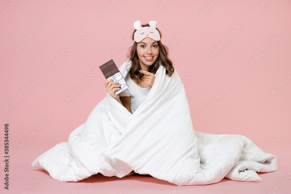 Smiling young woman in pajamas home wear sleep mask sitting pointing index finger on chocolate bar while resting at home isolated on pastel pink background studio portrait. Relax good mood concept. Smiling young woman in pajamas home wear sleep mask sitting pointing index finger on chocolate bar while resting at home isolated on pastel pink background studio portrait. Relax good mood concept.