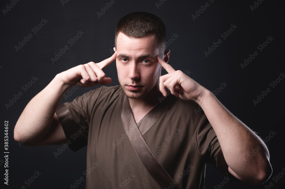 Portrait of a strong and large male masseur in special clothes with a finger at the temple reads your thoughts and knows all your diseases on a black background in the studio. Portrait of a strong and large male masseur in special clothes with a finger at the temple reads your thoughts and knows all your diseases on a black background in the studio.