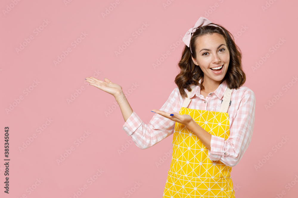 Cheerful funny young woman housewife in yellow apron pointing hands aside on mock up copy space while doing housework isolated on pastel pink colour background studio portrait. Housekeeping concept. Cheerful funny young woman housewife in yellow apron pointing hands aside on mock up copy space while doing housework isolated on pastel pink colour background studio portrait. Housekeeping concept.