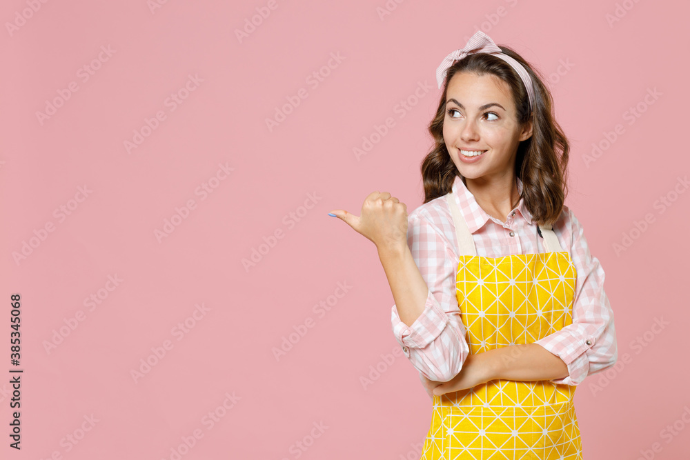 Smiling pretty young woman housewife in yellow apron pointing thumb aside on mock up copy space while doing housework isolated on pastel pink colour background studio portrait. Housekeeping concept. Smiling pretty young woman housewife in yellow apron pointing thumb aside on mock up copy space while doing housework isolated on pastel pink colour background studio portrait. Housekeeping concept.