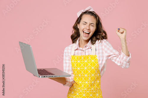 Obraz na płótnie Happy young woman housewife in yellow apron using laptop pc computer clenching fist like winner while doing housework isolated on pastel pink colour background studio portrait