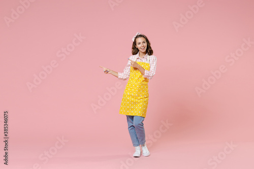Full length portrait of cheerful young woman housewife in yellow apron point index fingers aside on mock up copy space doing housework isolated on pastel pink background studio. Housekeeping concept.