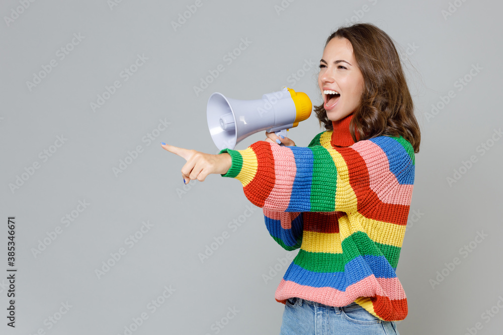 Excited cheerful young brunette woman 20s weaaring casual colorful knitted sweater standing screaming in megaphone pointing index finger aside isolated on grey colour background, studio portrait. Excited cheerful young brunette woman 20s weaaring casual colorful knitted sweater standing screaming in megaphone pointing index finger aside isolated on grey colour background, studio portrait.