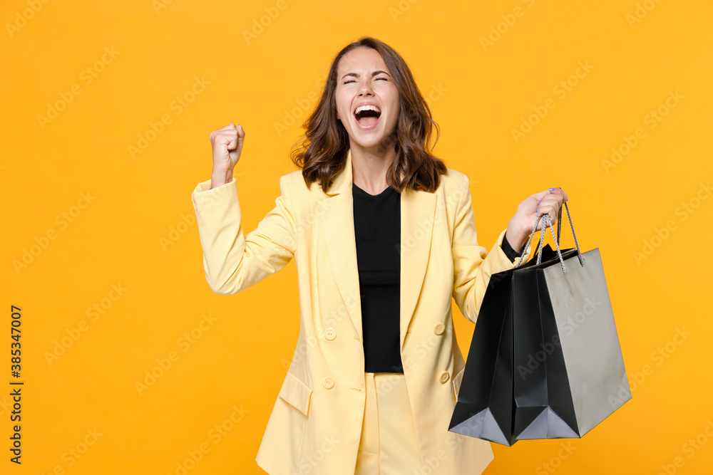 Overjoyed young woman in basic light suit jacket hold package bags with purchases after shopping clenching fists doing winner gesture isolated on yellow background studio portrait. Black friday sale. Overjoyed young woman in basic light suit jacket hold package bags with purchases after shopping clenching fists doing winner gesture isolated on yellow background studio portrait. Black friday sale.