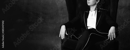 Black and white cropped photo of a woman in a black pantsuit sitting in a high chair in a comfortable position with one leg over the other. Cropped shot with dark gray background.