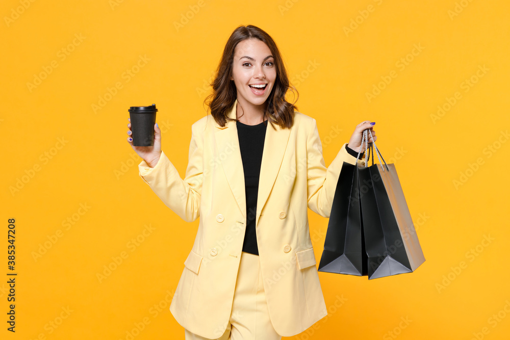 Excited young woman 20s in basic suit jacket hold package bags with purchases after shopping paper cup of coffee or tea looking camera isolated on yellow background studio portrait. Black friday sale. Excited young woman 20s in basic suit jacket hold package bags with purchases after shopping paper cup of coffee or tea looking camera isolated on yellow background studio portrait. Black friday sale.