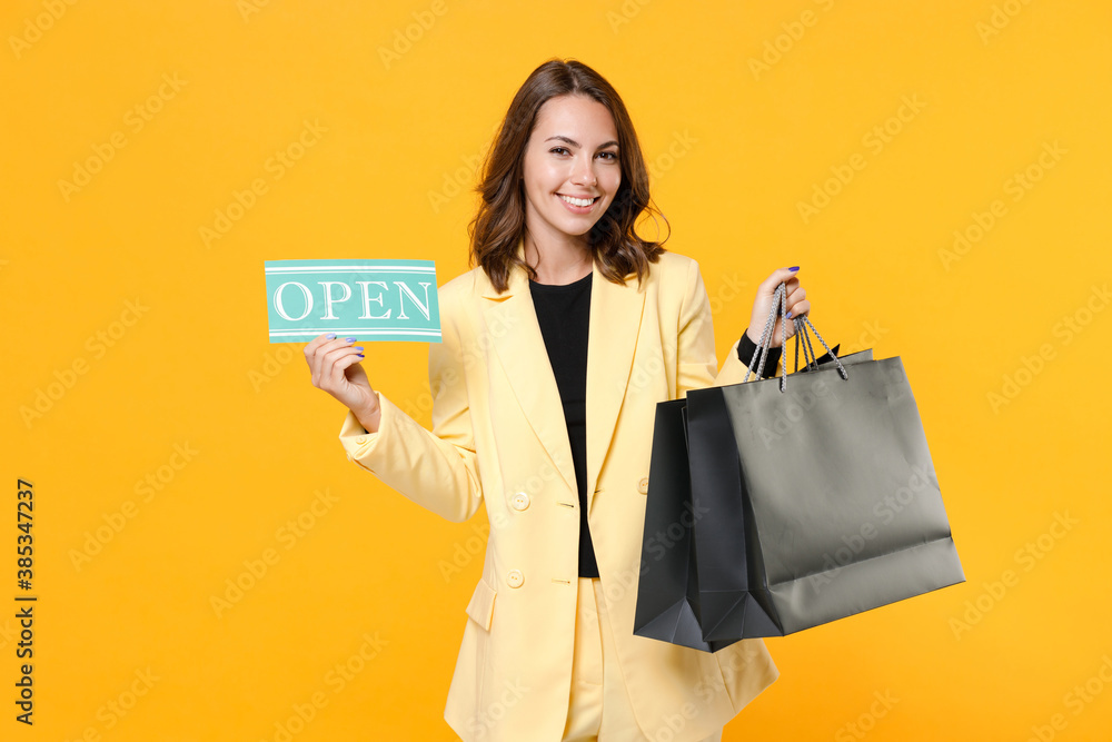 Smiling young brunette woman 20s wearing basic light suit jacket hold package bags with purchases after shopping sign with OPEN title isolated on yellow background, studio portrait. Black friday sale. Smiling young brunette woman 20s wearing basic light suit jacket hold package bags with purchases after shopping sign with OPEN title isolated on yellow background, studio portrait. Black friday sale.