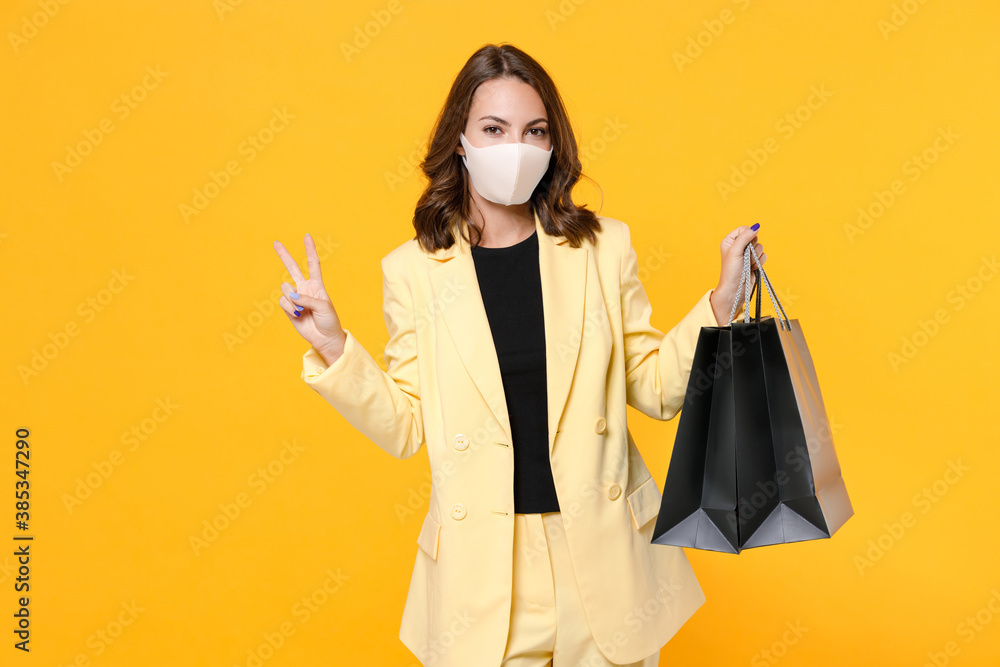 Young woman in jacket face mask to safe from coronavirus virus covid-19 hold package bags with purchases after shopping showing victory sign isolated on yellow background studio. Black friday sale. Young woman in jacket face mask to safe from coronavirus virus covid-19 hold package bags with purchases after shopping showing victory sign isolated on yellow background studio. Black friday sale.