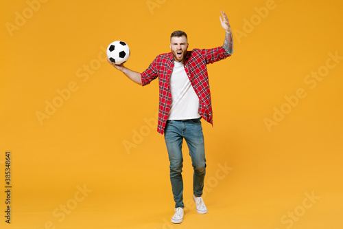 Full length portrait angry man football fan in red shirt cheer up support favorite team with soccer ball spreading hands screaming swearing isolated on yellow background. People sport leisure concept. © ViDi Studio