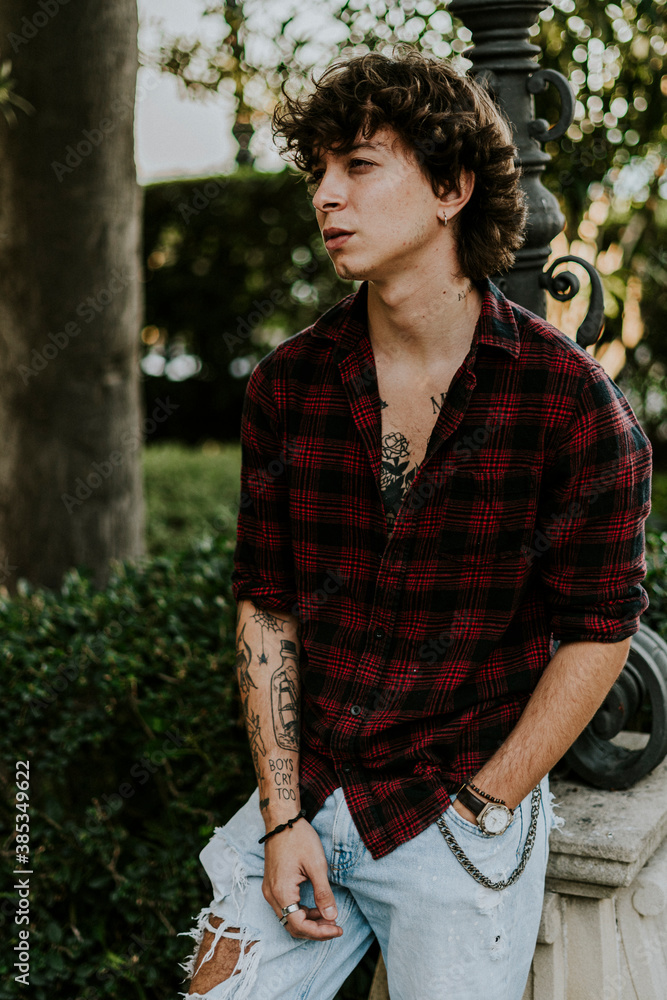 Hombre Joven moreno con tatuajes ,camisa roja de cuadros y vaquero azules  en parque de flores y arboles. Stock Photo | Adobe Stock