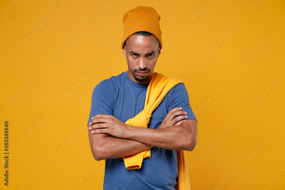 Displeased dissatisfied offended young african american man 20s wearing basic casual blue t-shirt hat holding hands crossed looking camera isolated on bright yellow colour background, studio portrait.
