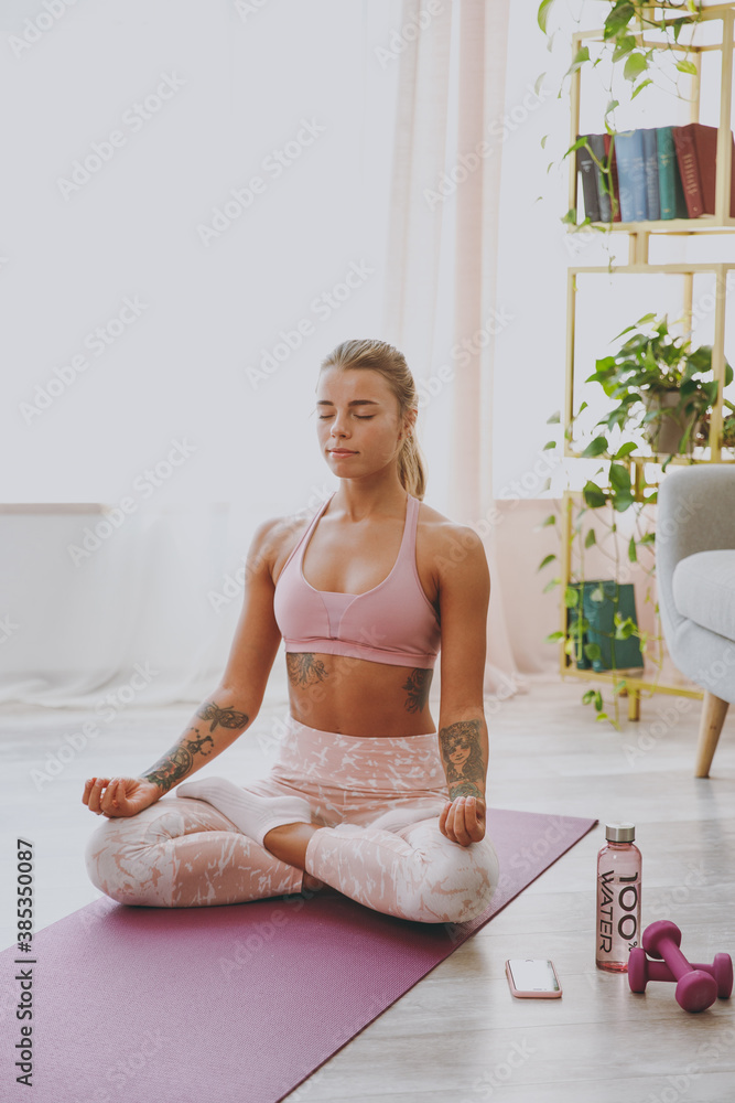 Beautiful attractive young strong sporty fitness woman wearing pink tracksuit doing yoga exercises sitting relaxing meditating trying to calm down keeping eyes closed stretching on mat at home indoor. Beautiful attractive young strong sporty fitness woman wearing pink tracksuit doing yoga exercises sitting relaxing meditating trying to calm down keeping eyes closed stretching on mat at home indoor.
