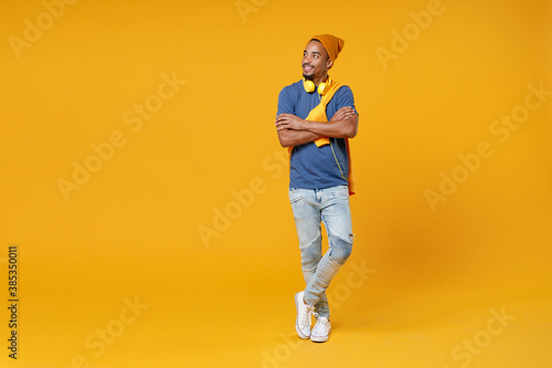 Full length of smiling attractive young african american man 20s wearing blue t-shirt hat standing holding hands crossed looking aside isolated on bright yellow colour background, studio portrait.