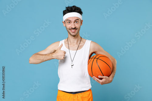 Smiling fitness man basketball player with thin skinny body sportsman in headband shirt shorts whistle hold ball showing thumb up isolated on blue background. Workout gym sport motivation concept. © ViDi Studio