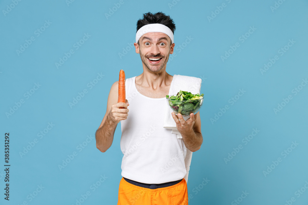 Excited young sporty fitness man with thin skinny body sportsman in headband shirt shorts hold vegetable salad in glass bowl carrot isolated on blue background. Workout gym sport motivation concept. Excited young sporty fitness man with thin skinny body sportsman in headband shirt shorts hold vegetable salad in glass bowl carrot isolated on blue background. Workout gym sport motivation concept.