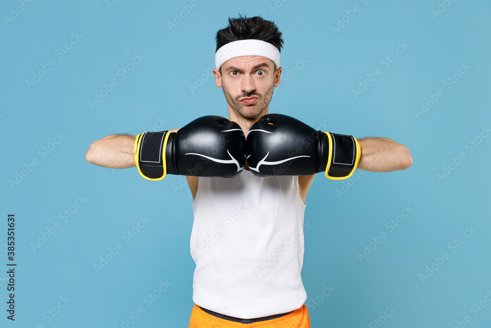Perplexed young strong sporty fitness man boxer with skinny body sportsman  in headband shirt shorts doing boxing exercises in gloves isolated on blue  background. Workout gym sport motivation concept. Stock Photo
