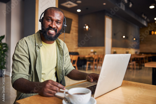 Joyful Afro American man in headphones using laptop in cafe