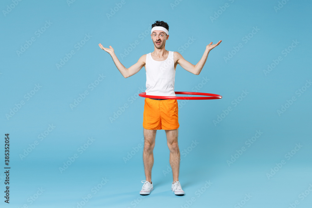 Full length portrait confused young man with skinny body sportsman in headband shirt shorts training with hula hoop spreading hands isolated on blue background. Workout gym sport motivation concept. Full length portrait confused young man with skinny body sportsman in headband shirt shorts training with hula hoop spreading hands isolated on blue background. Workout gym sport motivation concept.