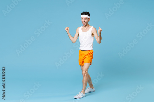 Full length portrait joyful young sporty fitness man with skinny body sportsman in white headband shirt shorts doing winner gesture isolated on blue background. Workout gym sport motivation concept.