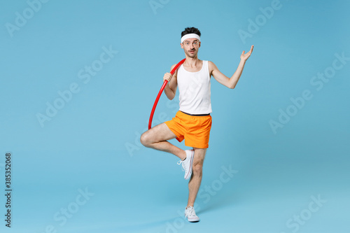 Full length portrait of puzzled young man with skinny body sportsman in headband shirt shorts training with hula hoop spreading hands isolated on blue background. Workout gym sport motivation concept.
