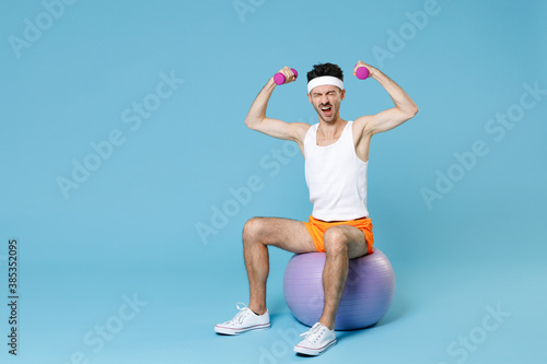 Full length portrait screaming man with skinny body sportsman in headband shirt shorts sit on fitball doing exercise with dumbbells isolated on blue background. Workout gym sport motivation concept.