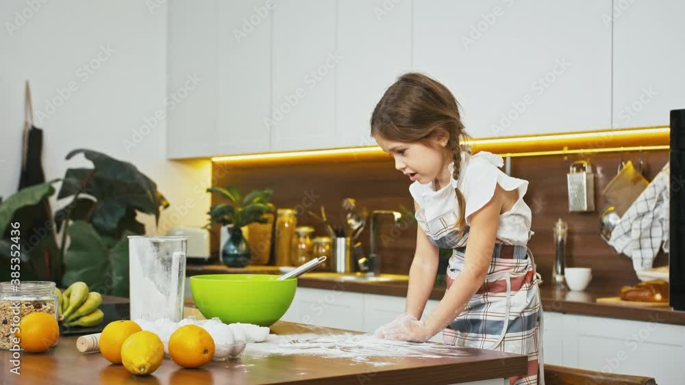 Little girl is playing hands with flour standing at a big table with ingredients for cooking on it, at modern kitchen Little girl is playing hands with flour standing at a big table with ingredients for cooking on it, at modern kitchen