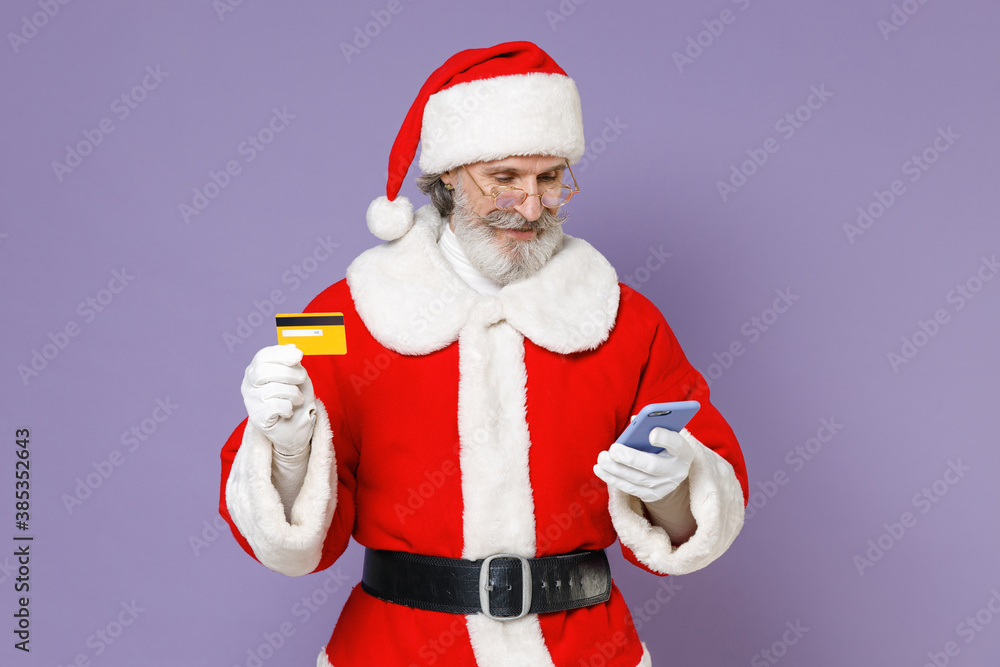 Elderly Santa Claus man in Christmas hat red suit coat white gloves glasses using mobile phone hold credit bank card isolated on violet background. Happy New Year celebration merry holiday concept. Elderly Santa Claus man in Christmas hat red suit coat white gloves glasses using mobile phone hold credit bank card isolated on violet background. Happy New Year celebration merry holiday concept.