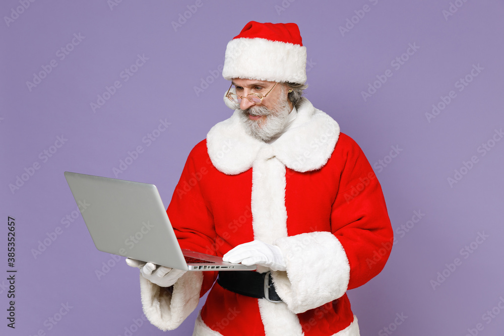 Naklejka premium Elderly gray-haired Santa Claus man in Christmas hat red suit coat white gloves glasses working on laptop pc computer isolated on violet background. Happy New Year celebration merry holiday concept.