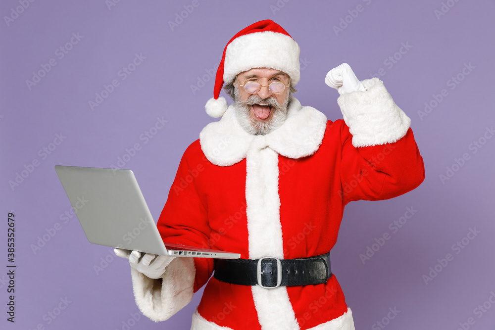 Blinking Santa Claus man in Christmas hat red coat gloves glasses working on laptop pc computer doing winner gesture isolated on violet background. Happy New Year celebration merry holiday concept. Blinking Santa Claus man in Christmas hat red coat gloves glasses working on laptop pc computer doing winner gesture isolated on violet background. Happy New Year celebration merry holiday concept.