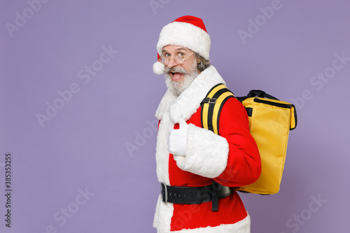 Side view of excited Santa Claus man in Christmas hat red suit coat glasses thermal food bag backpack showing thumb up isolated on violet background. Happy New Year celebration merry holiday concept.