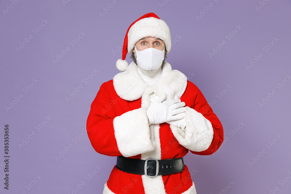 Santa Claus man in Christmas hat red coat face mask to safe from coronavirus virus covid-19 hold hands folded on heart isolated on violet background. Happy New Year celebration merry holiday concept. Santa Claus man in Christmas hat red coat face mask to safe from coronavirus virus covid-19 hold hands folded on heart isolated on violet background. Happy New Year celebration merry holiday concept.