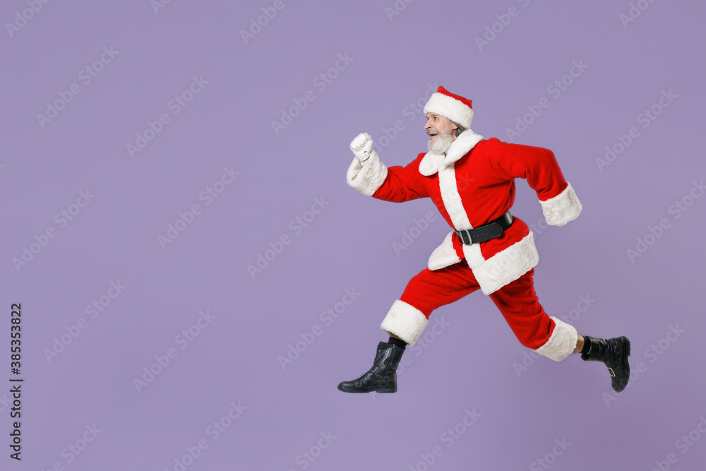 Full length side view portrait of funny Santa Claus man in Christmas hat red suit coat gloves jumping like running isolated on violet background. Happy New Year celebration merry holiday concept. Full length side view portrait of funny Santa Claus man in Christmas hat red suit coat gloves jumping like running isolated on violet background. Happy New Year celebration merry holiday concept.