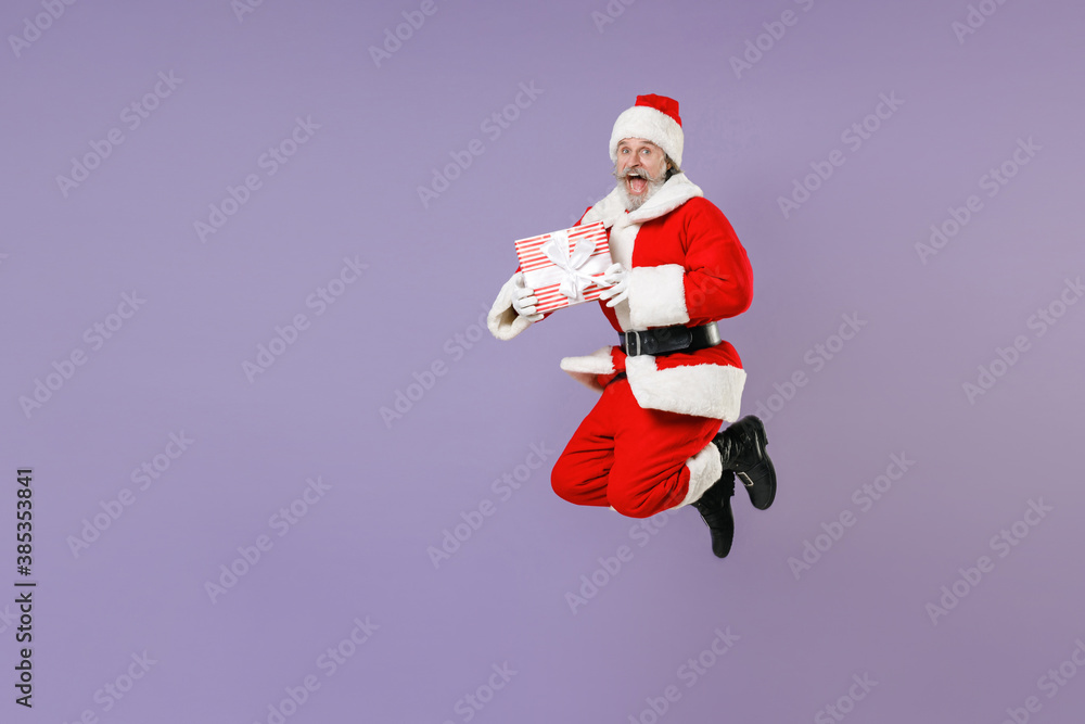 Full length portrait of excited Santa Claus man in Christmas hat red coat jumping hold present box with gift ribbon bow isolated on violet background. Happy New Year celebration merry holiday concept. Full length portrait of excited Santa Claus man in Christmas hat red coat jumping hold present box with gift ribbon bow isolated on violet background. Happy New Year celebration merry holiday concept.