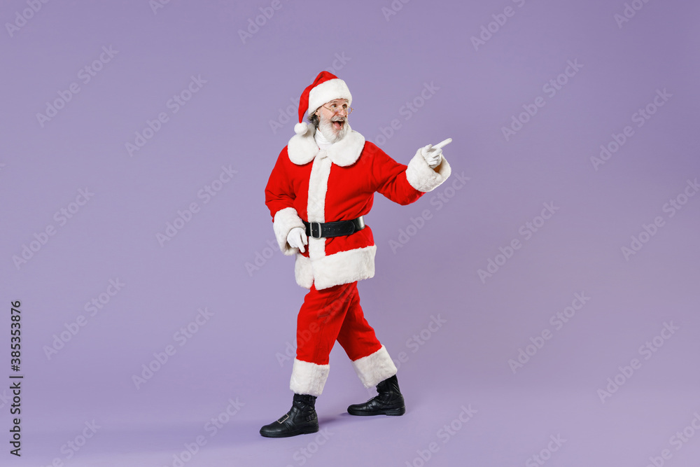 Full length side view portrait of excited Santa Claus man in Christmas hat red coat suit pointing index finger aside isolated on violet background. Happy New Year celebration merry holiday concept.