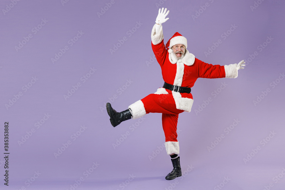 Full length portrait excited Santa Claus man in Christmas hat red suit coat glasses dancing spreading hands rising leg isolated on violet background. Happy New Year celebration merry holiday concept. Full length portrait excited Santa Claus man in Christmas hat red suit coat glasses dancing spreading hands rising leg isolated on violet background. Happy New Year celebration merry holiday concept.