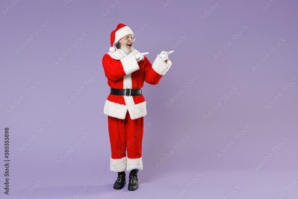 Fototapeta premium Full length portrait of Santa Claus man in Christmas hat red suit coat white gloves glasses point index fingers aside isolated on violet background. Happy New Year celebration merry holiday concept.