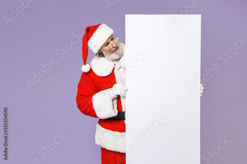 Cheerful Santa Claus man in Christmas hat red coat suit gloves glasses hold big white empty blank billboard isolated on violet background in studio. Happy New Year celebration merry holiday concept. © ViDi Studio