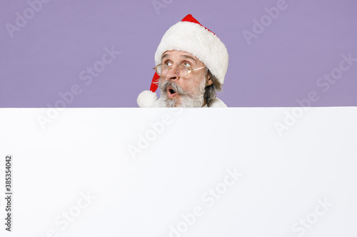Shocked amazed Santa Claus man in Christmas hat red coat gloves glasses hold big white empty blank billboard isolated on violet background studio. Happy New Year celebration merry holiday concept.