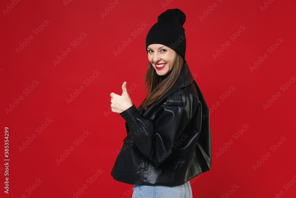 Side view of smiling beautiful young brunette woman 20s wearing casual black leather jacket white t-shirt showing thumb up looking camera isolated on bright red colour background, studio portrait.