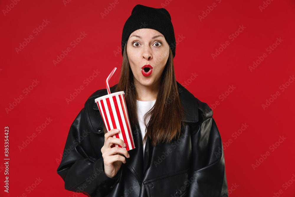 Shocked amazed young brunette woman 20s wearing casual basic black leather jacket white t-shirt hat hold cup of cola or soda keeping mouth open isolated on bright red background studio portrait. Shocked amazed young brunette woman 20s wearing casual basic black leather jacket white t-shirt hat hold cup of cola or soda keeping mouth open isolated on bright red background studio portrait.