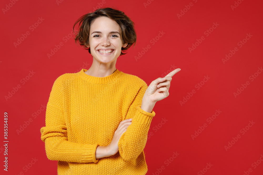 Smiling cheerful beautiful young brunette woman 20s wearing casual yellow sweater standing pointing index finger aside on mock up copy space isolated on bright red colour background studio portrait. Smiling cheerful beautiful young brunette woman 20s wearing casual yellow sweater standing pointing index finger aside on mock up copy space isolated on bright red colour background studio portrait.