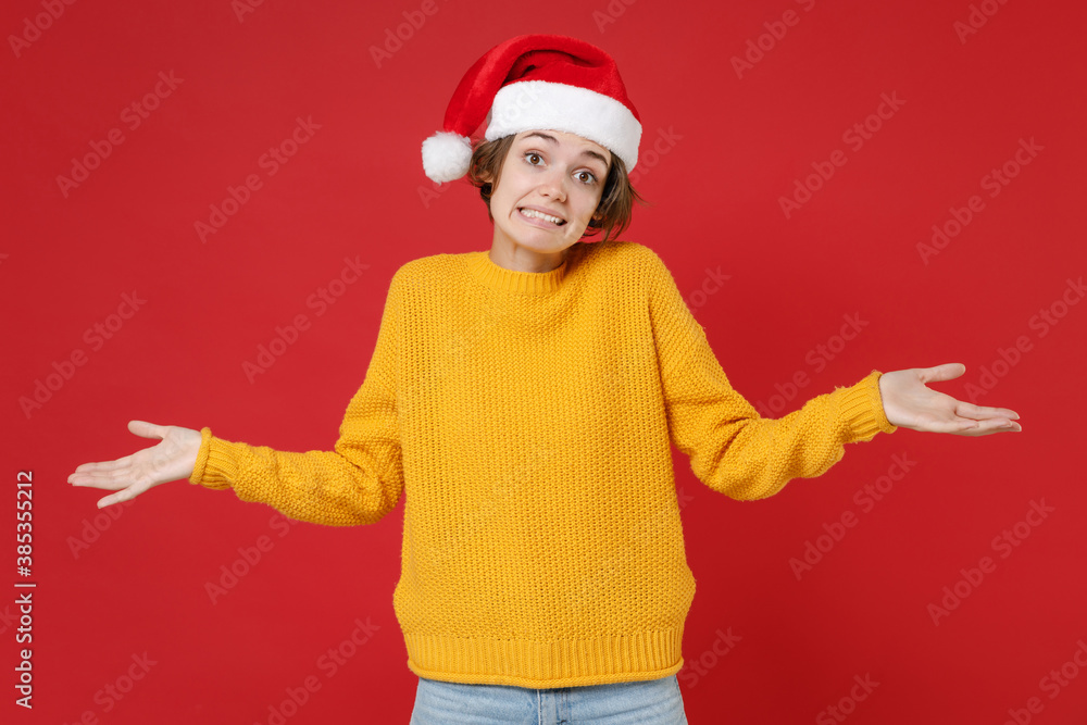 Confused puzzled young brunette Santa woman wearing casual yellow sweater Christmas hat spreading hands isolated on red background studio portrait. Happy New Year celebration merry holiday concept.