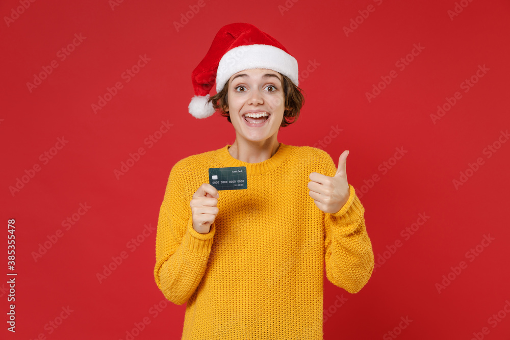 Excited young Santa woman in casual yellow sweater Christmas hat hold credit bank card showing thumb up isolated on red background studio portrait. Happy New Year celebration merry holiday concept. Excited young Santa woman in casual yellow sweater Christmas hat hold credit bank card showing thumb up isolated on red background studio portrait. Happy New Year celebration merry holiday concept.