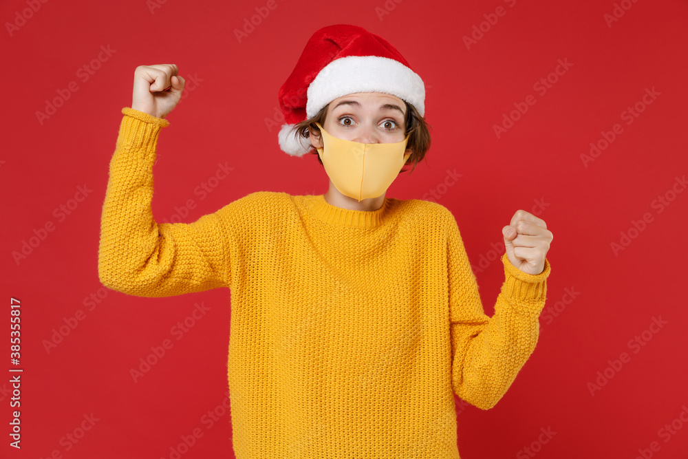 Excited Santa woman in Christmas hat face mask safe from coronavirus virus covid-19 clenching fists doing winner gesture isolated on red background. Happy New Year celebration merry holiday concept. Excited Santa woman in Christmas hat face mask safe from coronavirus virus covid-19 clenching fists doing winner gesture isolated on red background. Happy New Year celebration merry holiday concept.