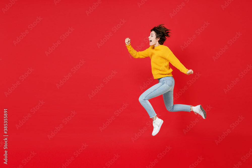 Full length side view of excited surprised cheerful funny young brunette woman 20s wearing basic casual yellow sweater jumping like running isolated on bright red colour background studio portrait.