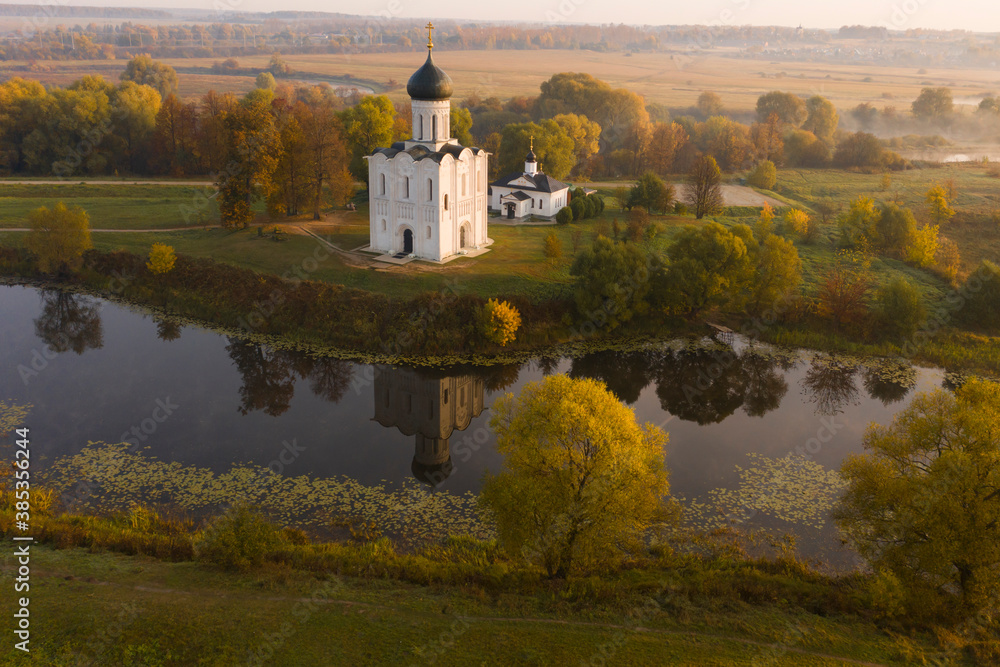 Aerial shot of a Russian Orthodox church above a misty field lit by the rising sun. A pond near the church, a river in the background. No people.